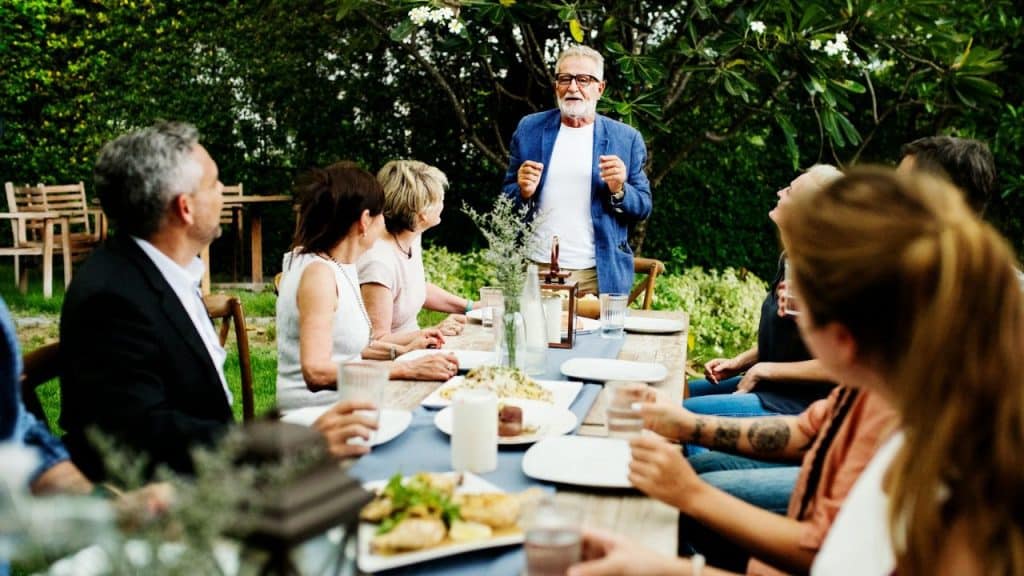 A man in a blue jacket stands and speaks to people seated around a long table outdoors.