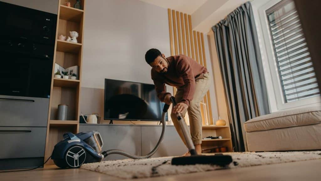 A man vacuums a patterned rug in a living room, looking towards the camera.