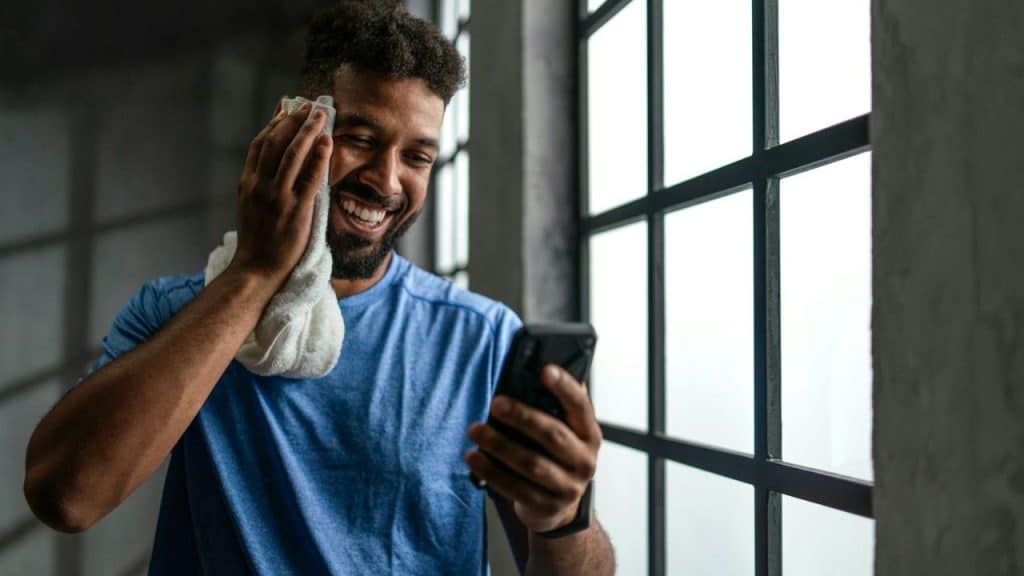 A smiling, bearded man in a blue shirt wipes his face with a towel and looks at his phone.