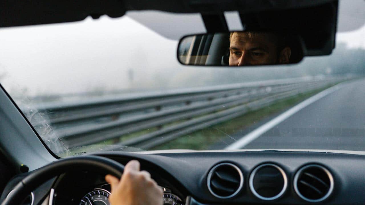 A person driving a car and is seen through the rearview mirror.