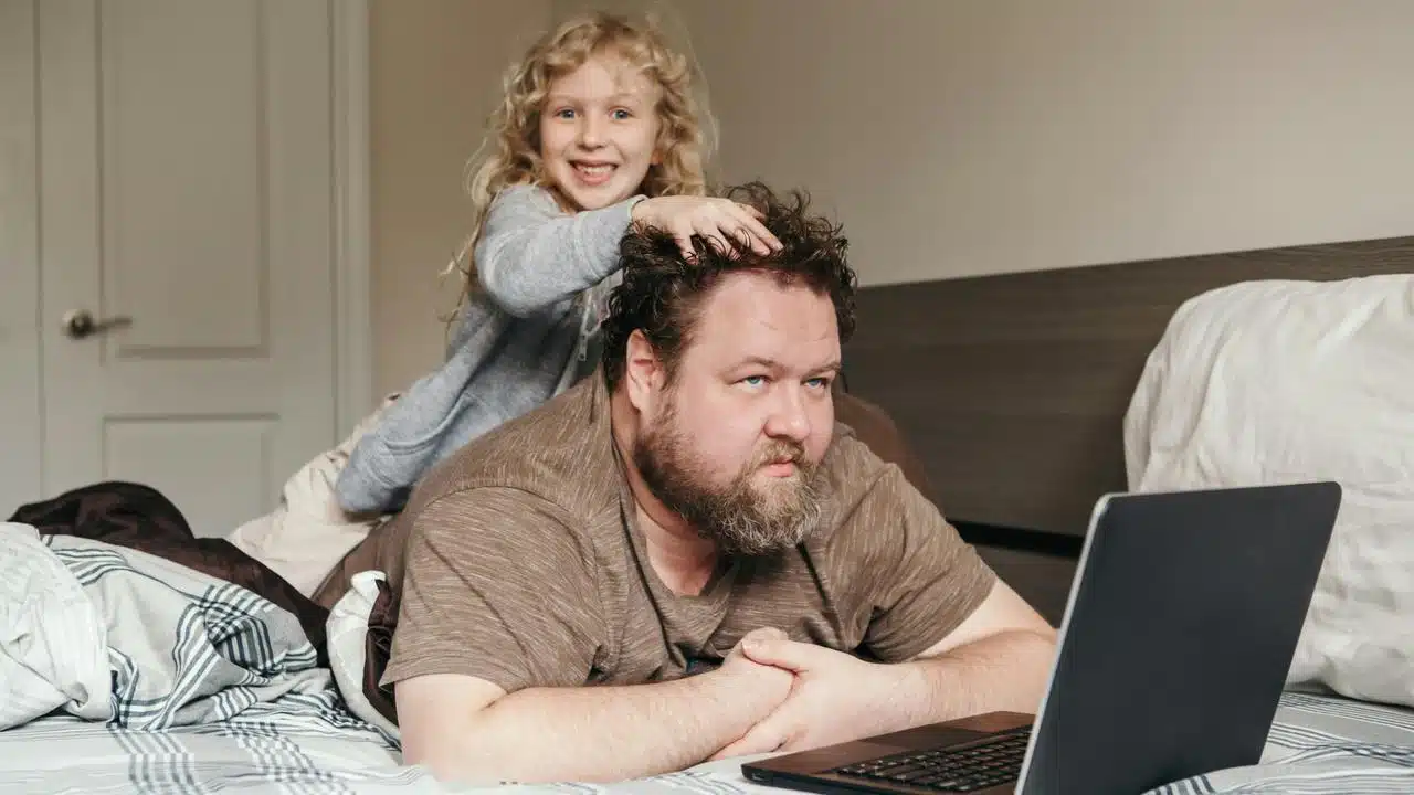 A man using a laptop while a child plays with his hair.