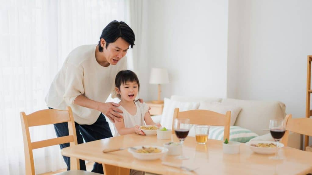 A man stands over a young girl seated at a dining table, holding a bowl of food.