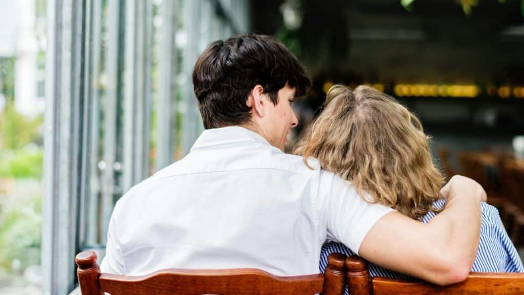 A man in a white shirt has his arm around a person with wavy hair, seen from behind.