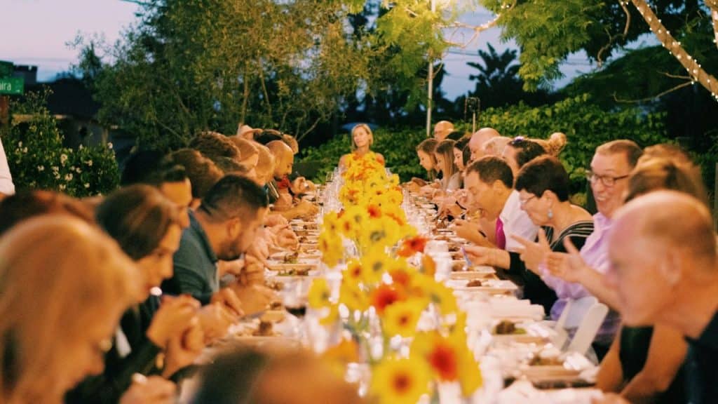 A large group of people are seated at a long table outdoors, adorned with yellow flowers.