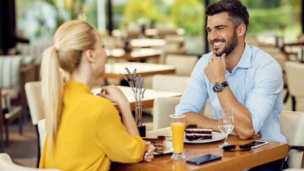 A smiling man with a beard sits across from a blonde woman at a table with food and drinks.