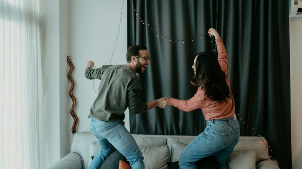 A man and a woman in jeans and long-sleeved shirts dance on a couch.
