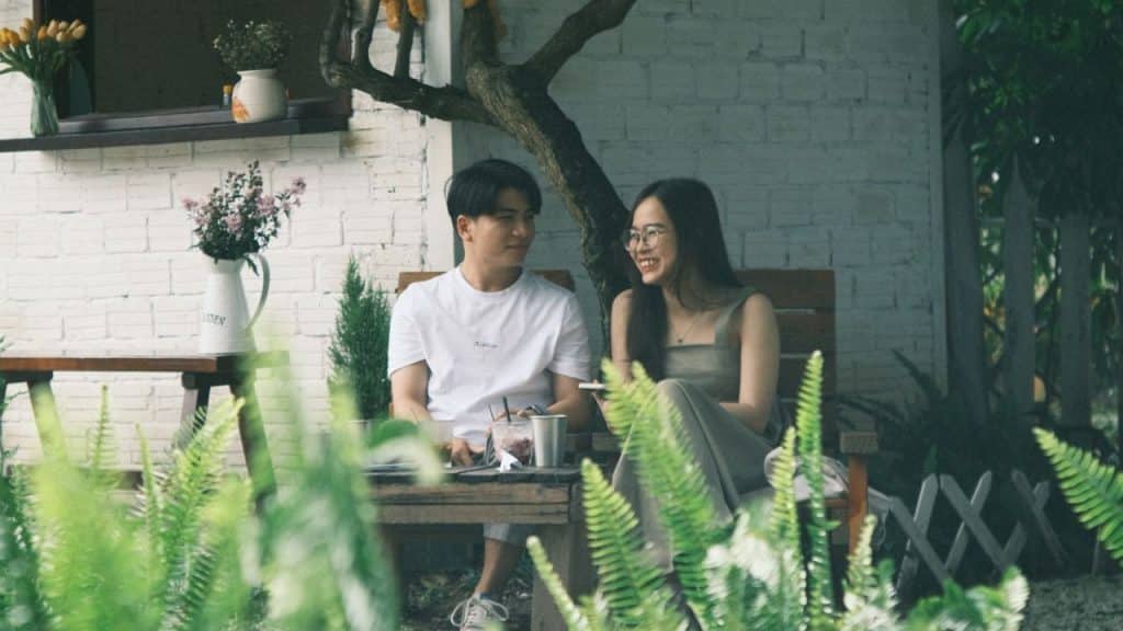 A man and woman sit smiling at a table outdoors, with foliage in the foreground.
