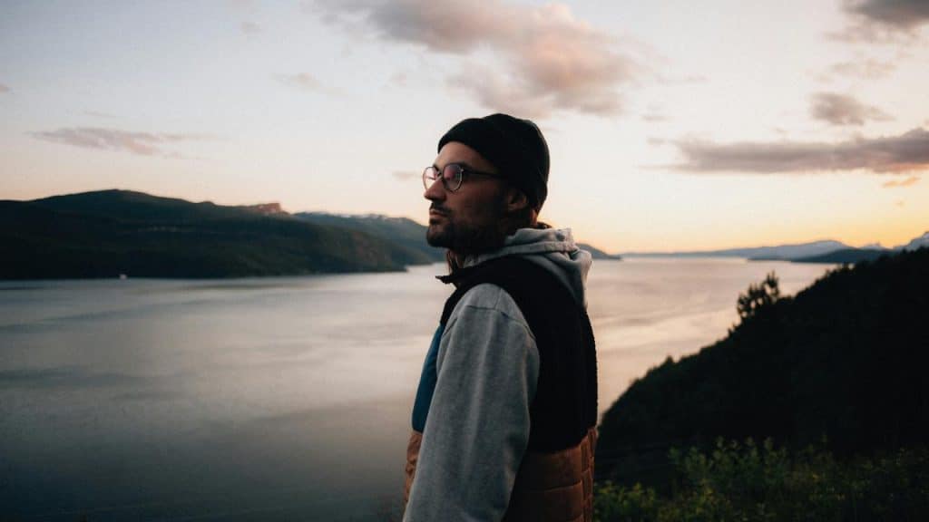 A man wearing glasses and a beanie standing by a lake at sunset.