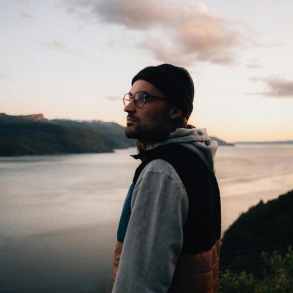 A man wearing glasses and a beanie standing by a lake at sunset.