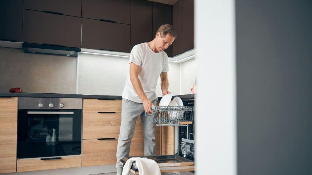 A man in a white t-shirt and gray sweatpants loads dishes into a dishwasher.