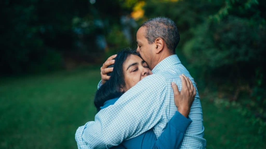 A man embraces a woman outdoors, kissing her on the forehead.
