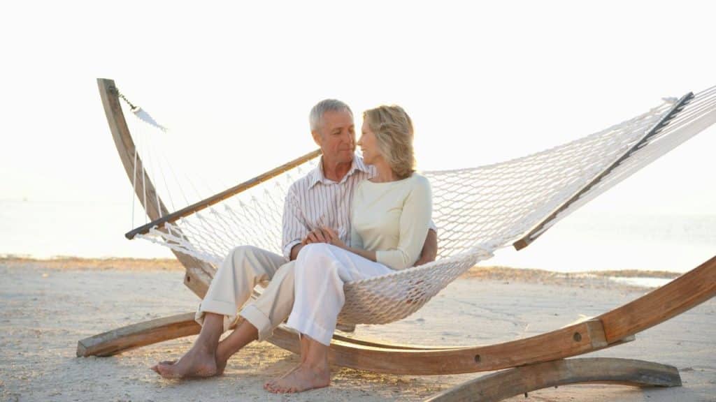 A man and woman sit in a hammock on a beach, looking at each other.