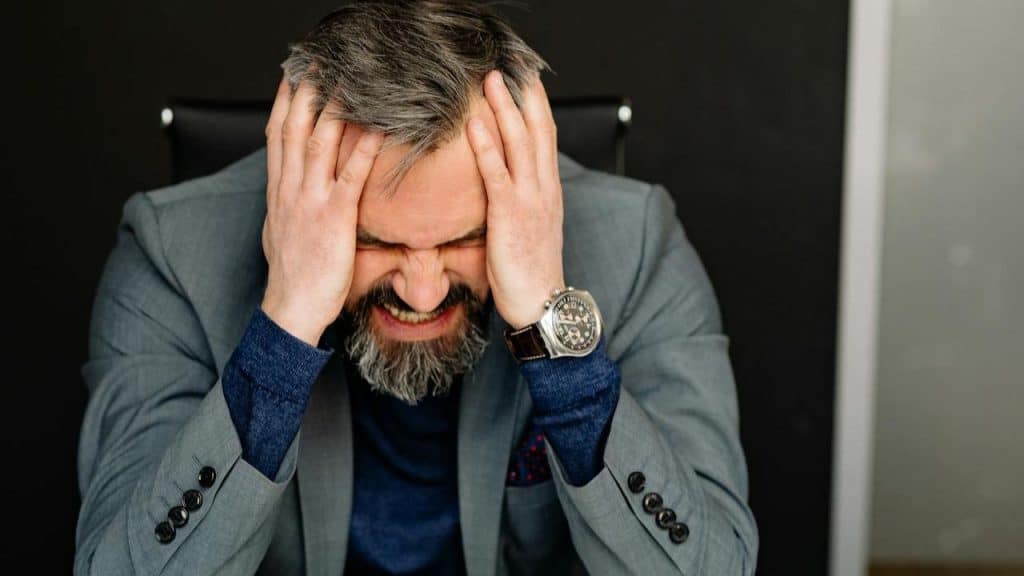 A man with a gray beard and gray hair clutches his head in frustration.