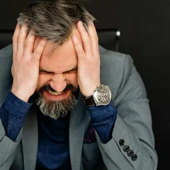 A man with a gray beard and gray hair clutches his head in frustration.