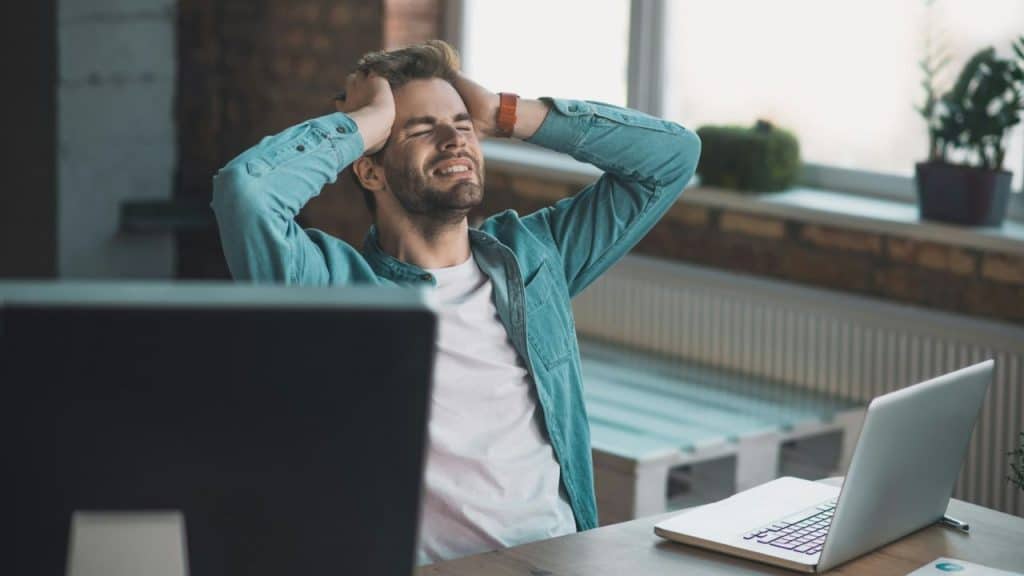 A man with a beard and a light blue shirt holds his head in his hands, looking frustrated.