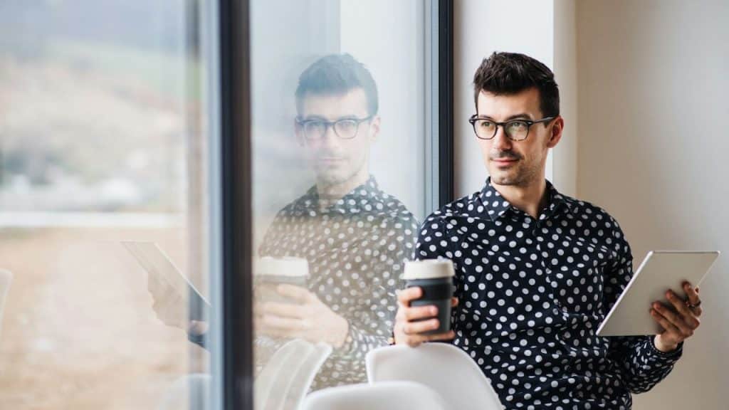 A man in a polka dot shirt holds a coffee cup and tablet, looking out a window.
