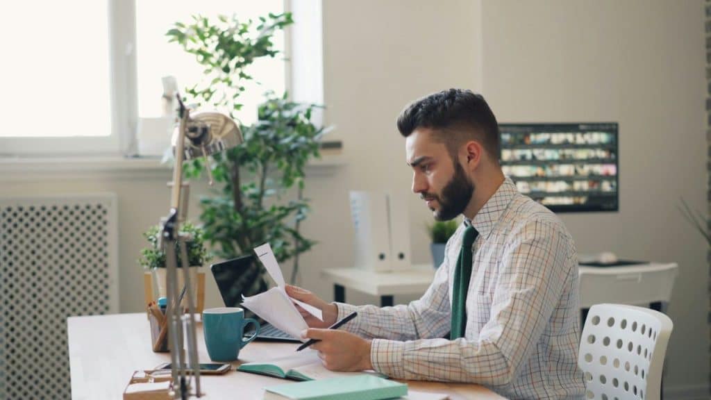 A man with a beard sits at a desk, looking at papers.