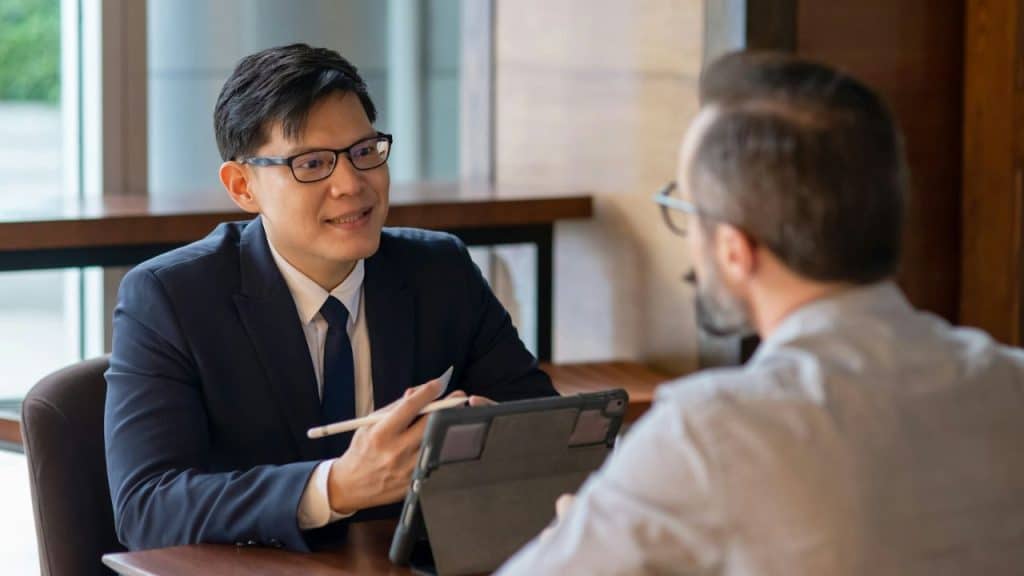 A man in a suit and glasses points at a tablet while talking to another person.
