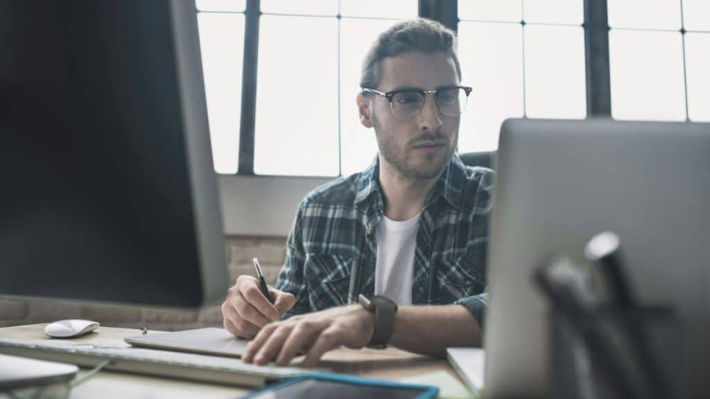A man wearing glasses and a plaid shirt works at a computer, writing on a clipboard.