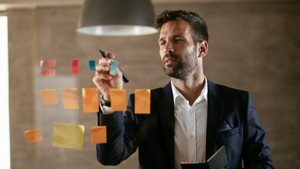 A man in a suit writes on a clear board covered with colorful sticky notes.