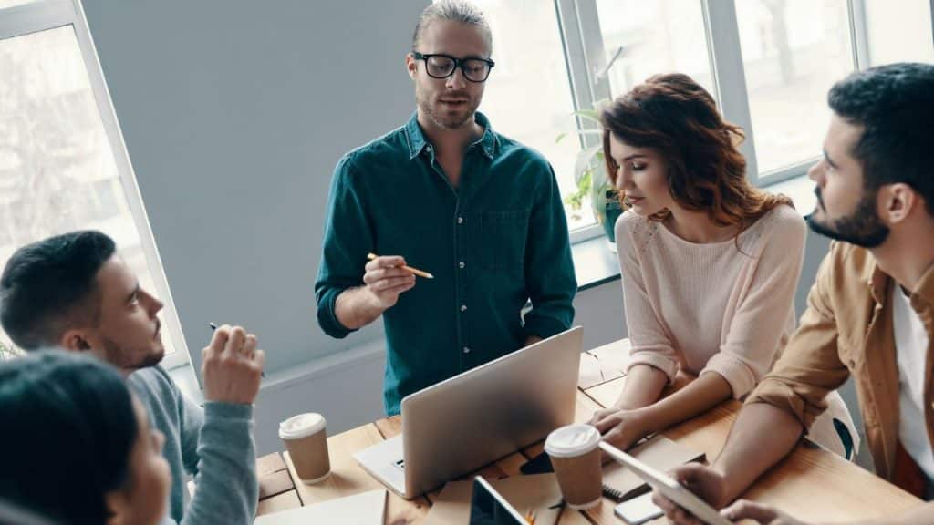 A man with a bun and glasses stands, leading a discussion with three other people.