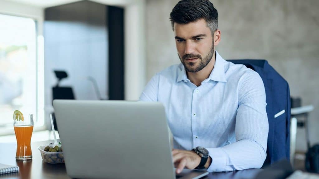 A man with a beard sits at a desk, looking at a laptop.