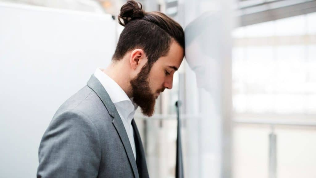 A man with a beard and hair bun leans his head against a white wall.
