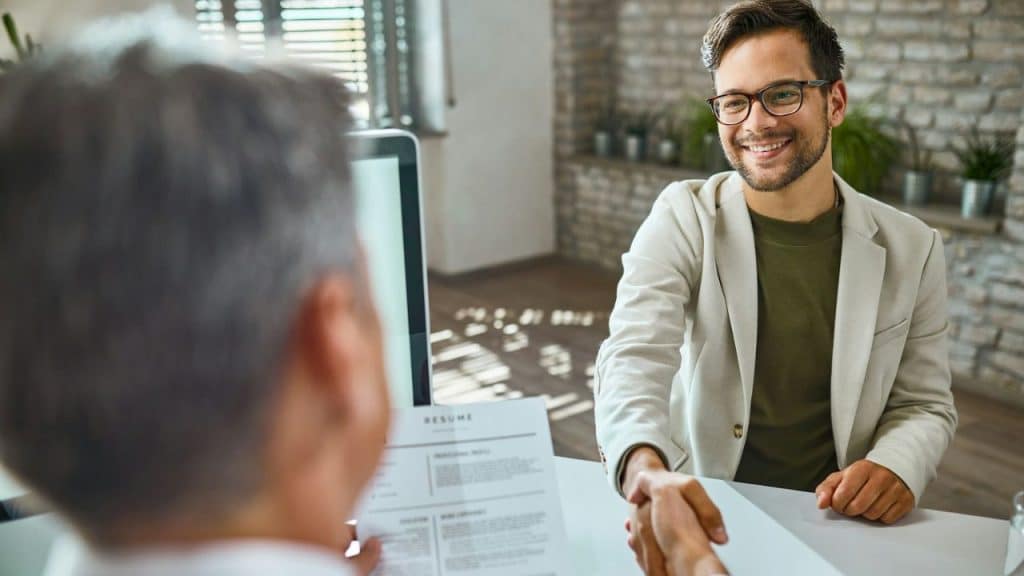 A smiling man in glasses and a light jacket shakes hands with someone across a desk.