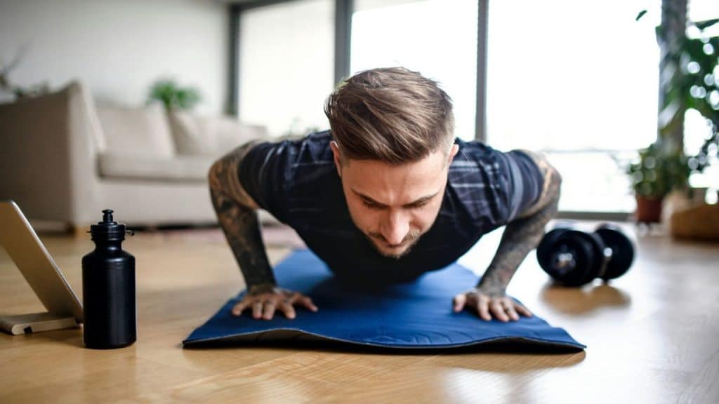 A man with tattoos on his arms does a push-up on a blue mat in a living room.