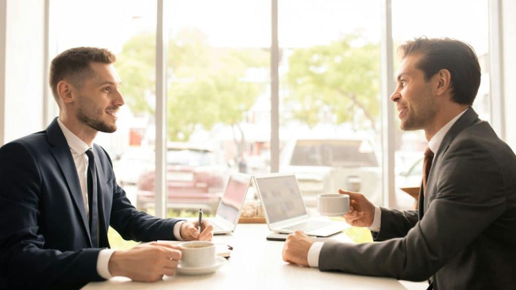 Two men in suits sit at a table by a window, talking and drinking coffee.