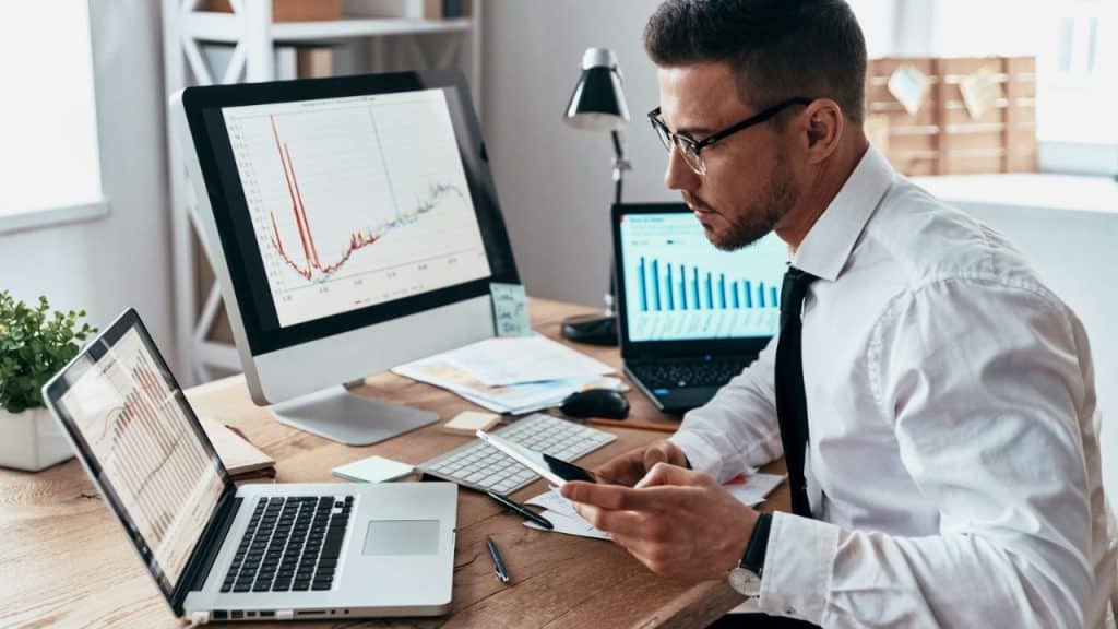 A man in glasses and a tie works at a desk with multiple screens displaying graphs.