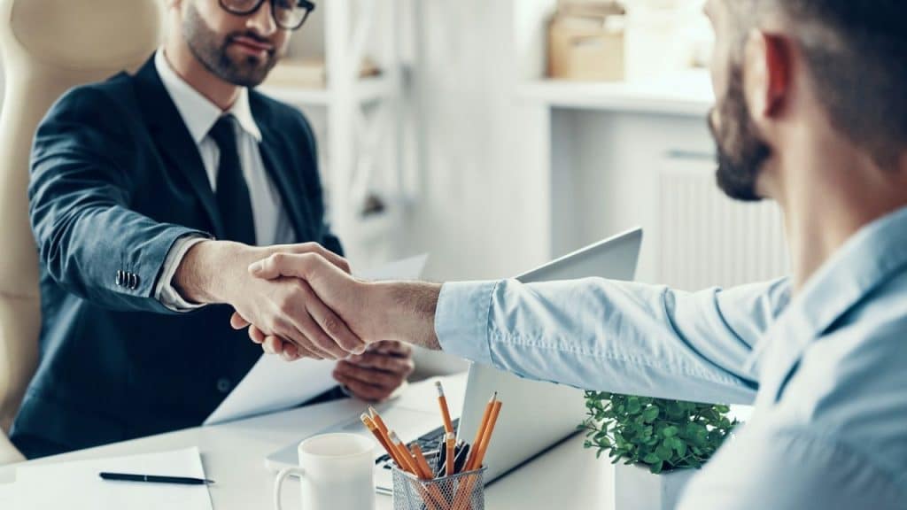 A man in a suit shakes hands with another man across a desk.