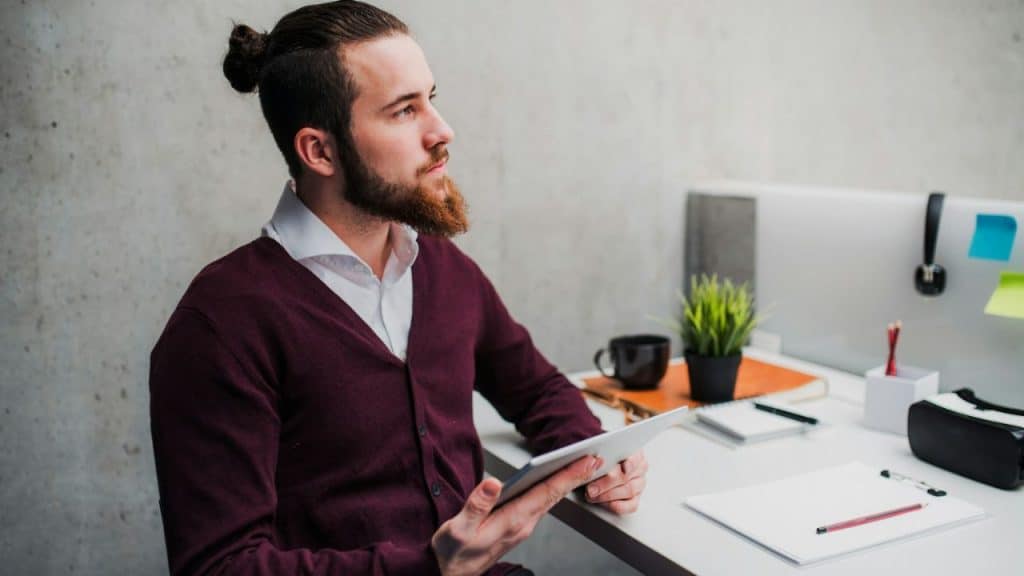 A man with a beard and hair bun holds a tablet, looking thoughtfully to the side at his desk.