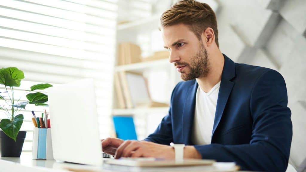 A man with a beard, wearing a blue blazer, looks intently at a laptop.
