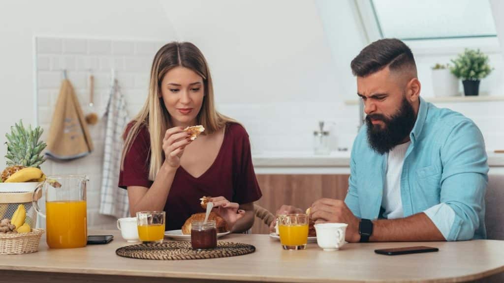 A woman eating breakfast while a man sitting beside her makes a disgusted face.