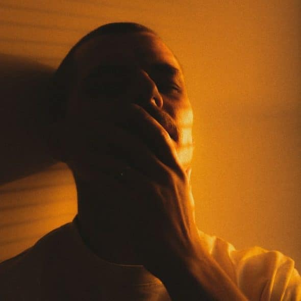 A man sits in a dimly lit room, covering his mouth with his hand.