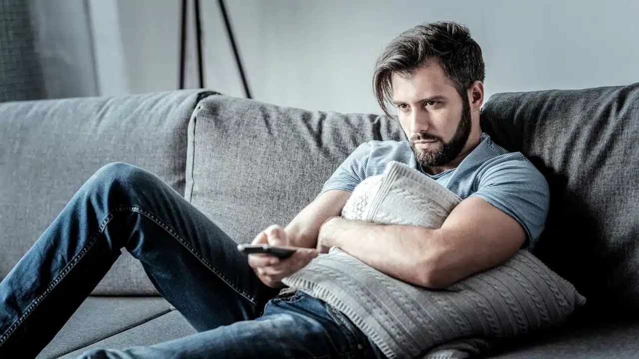 A man with a beard sits on a gray couch, holding a pillow and a remote.