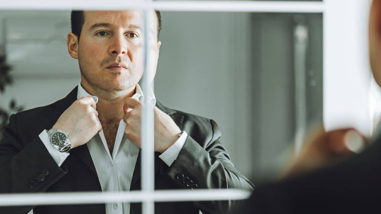 A man in a suit looks in a mirror, adjusting the collar of his white shirt.