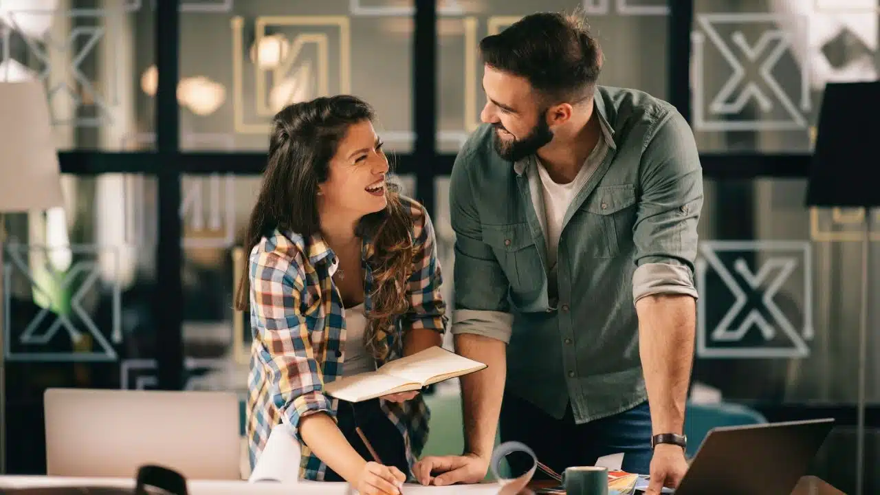 A happy man and woman smile at each other while working together at a desk.