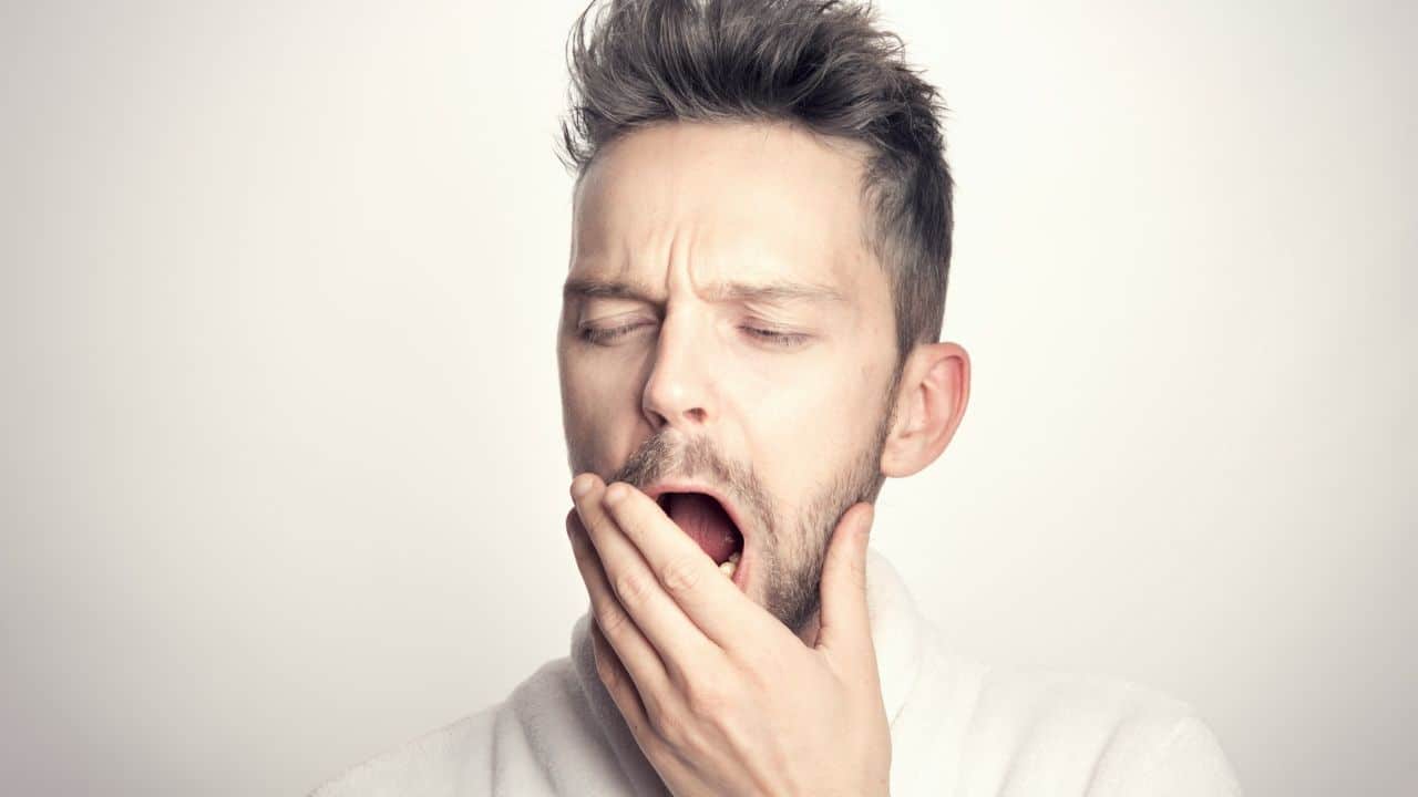 A man with messy hair yawns, covering his mouth with his hand.