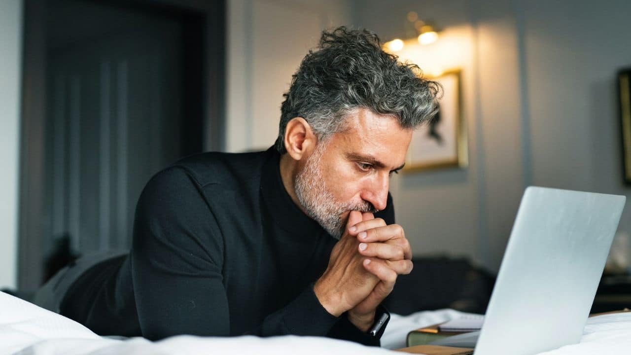 A man with salt and pepper hair is lying on a bed looking at a laptop.