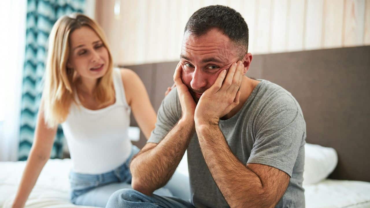 A frustrated man with his hands on his face looks at the camera while a woman is out of focus in the background.