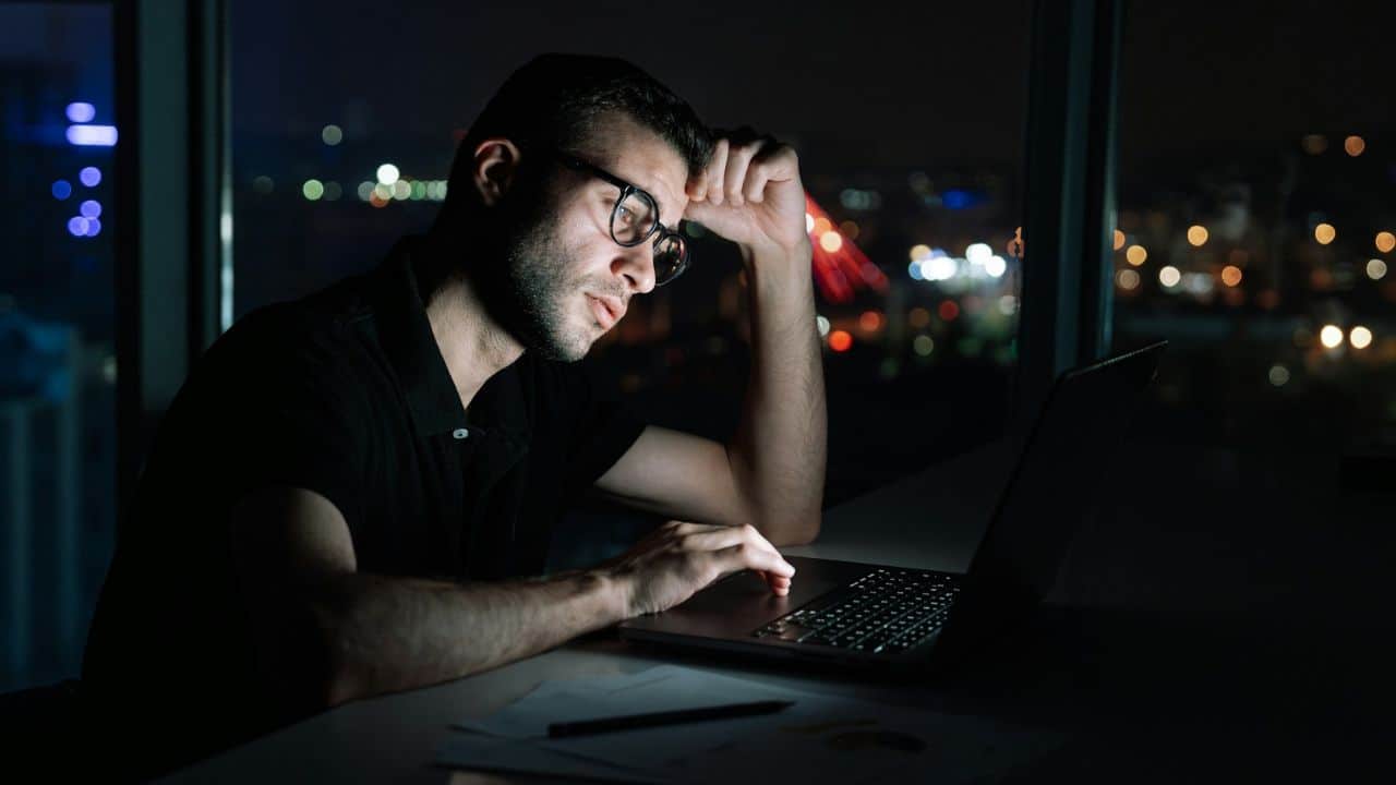 A tired man in glasses works on a laptop at night, with a blurry city view behind him.