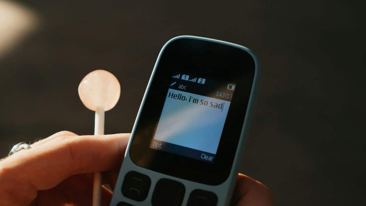 A hand holds an old mobile phone with a text message and a lollipop.