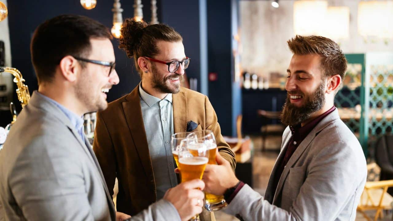 Three well-dressed men smile and toast with glasses of beer in a bar.