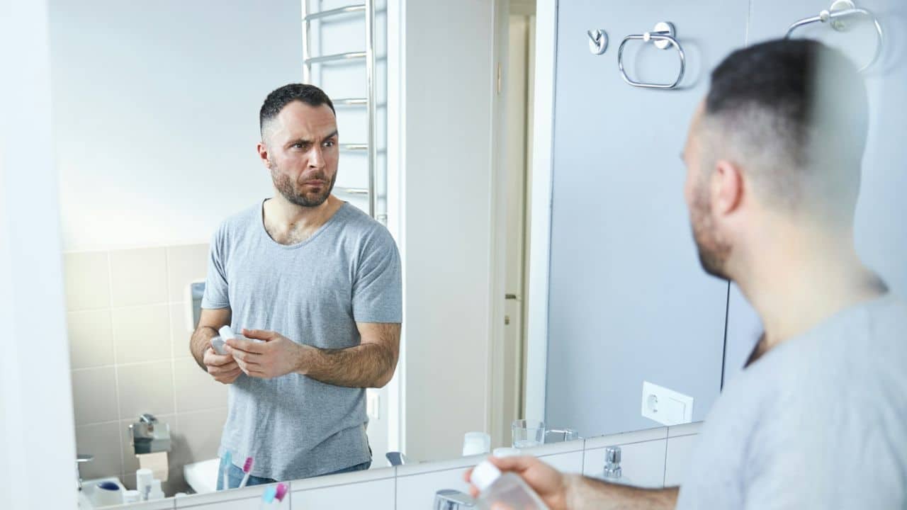 A man with a worried expression looks at his reflection in a bathroom mirror.