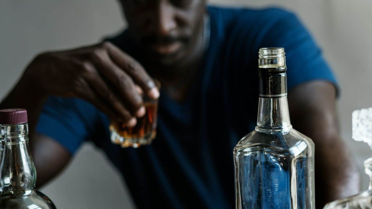 A blurred man holds a glass of whiskey, with bottles of liquor in the foreground.