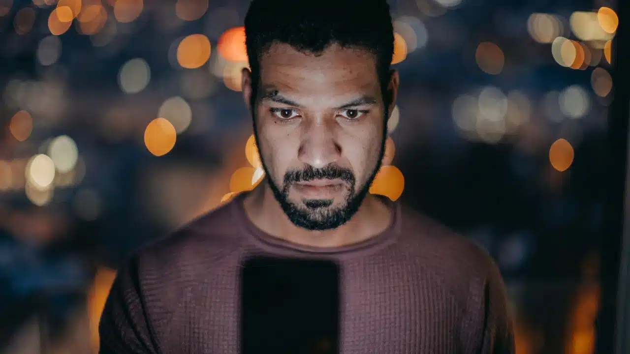A serious man looks intently at his phone at night, with city lights in the background.