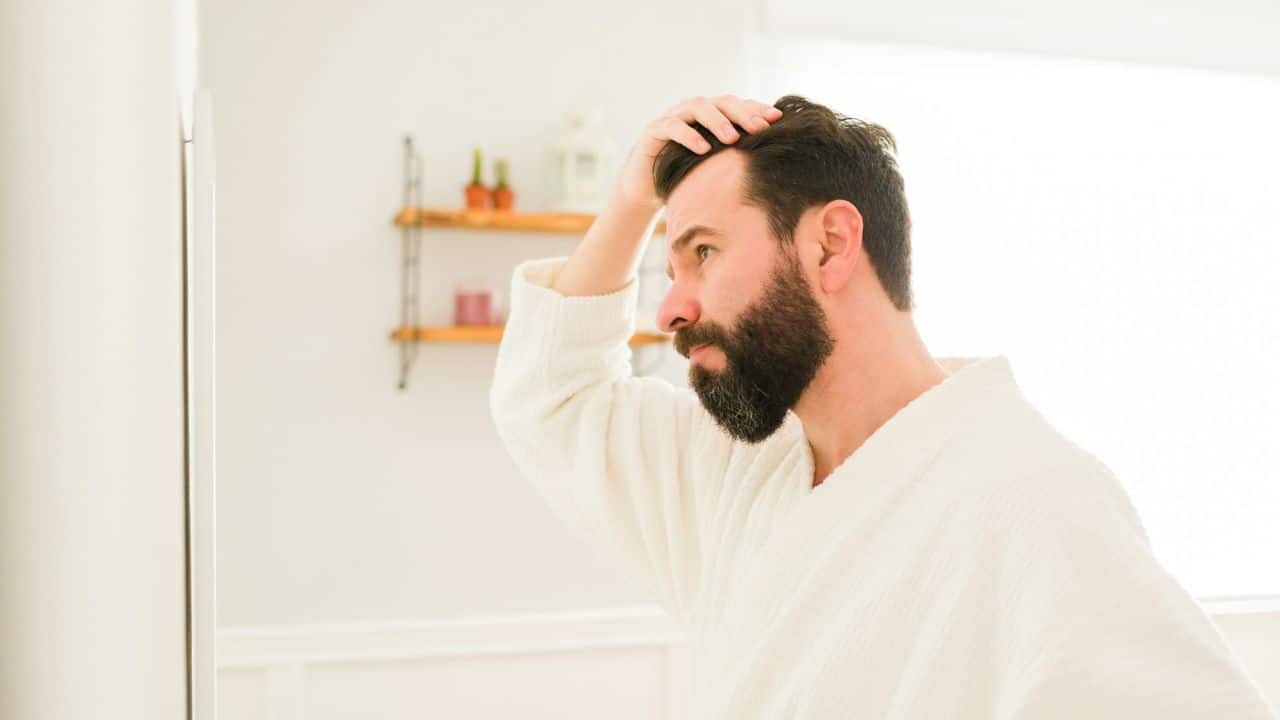 A man with a beard looks into a mirror, checking his hair and scalp.