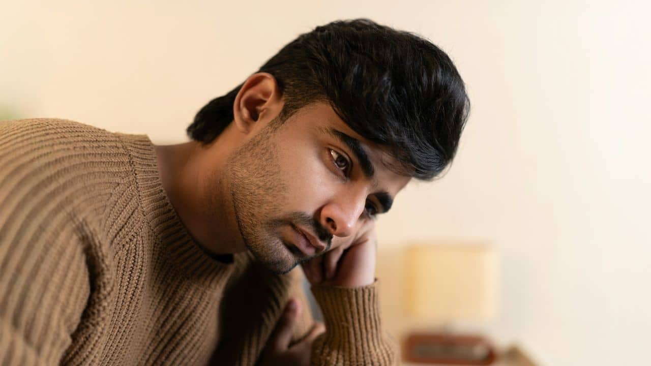 A thoughtful man in a brown sweater looks down with a serious expression.
